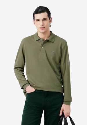 Jeune homme aux cheveux foncés coiffés en arrière porte un polo à manches longues vert olive et un pantalon foncé, tenant un sac noir dans une main.