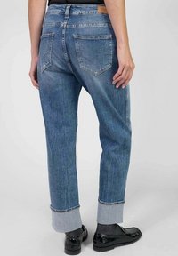Persona que lleva puestos unos pantalones de mezclilla azules con puños, calcetines negros y mocasines negros pulidos, mostrada desde atrás contra un fondo liso.