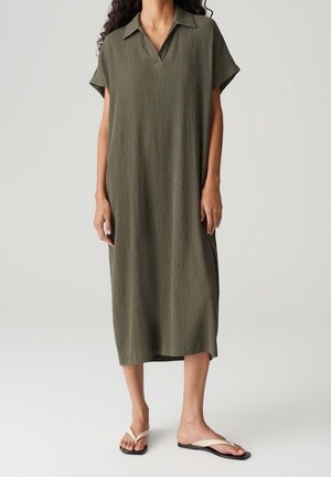 Femme portant une robe midi ample vert olive avec manches courtes et col, associée à des sandales plates blanches sur un fond uni.