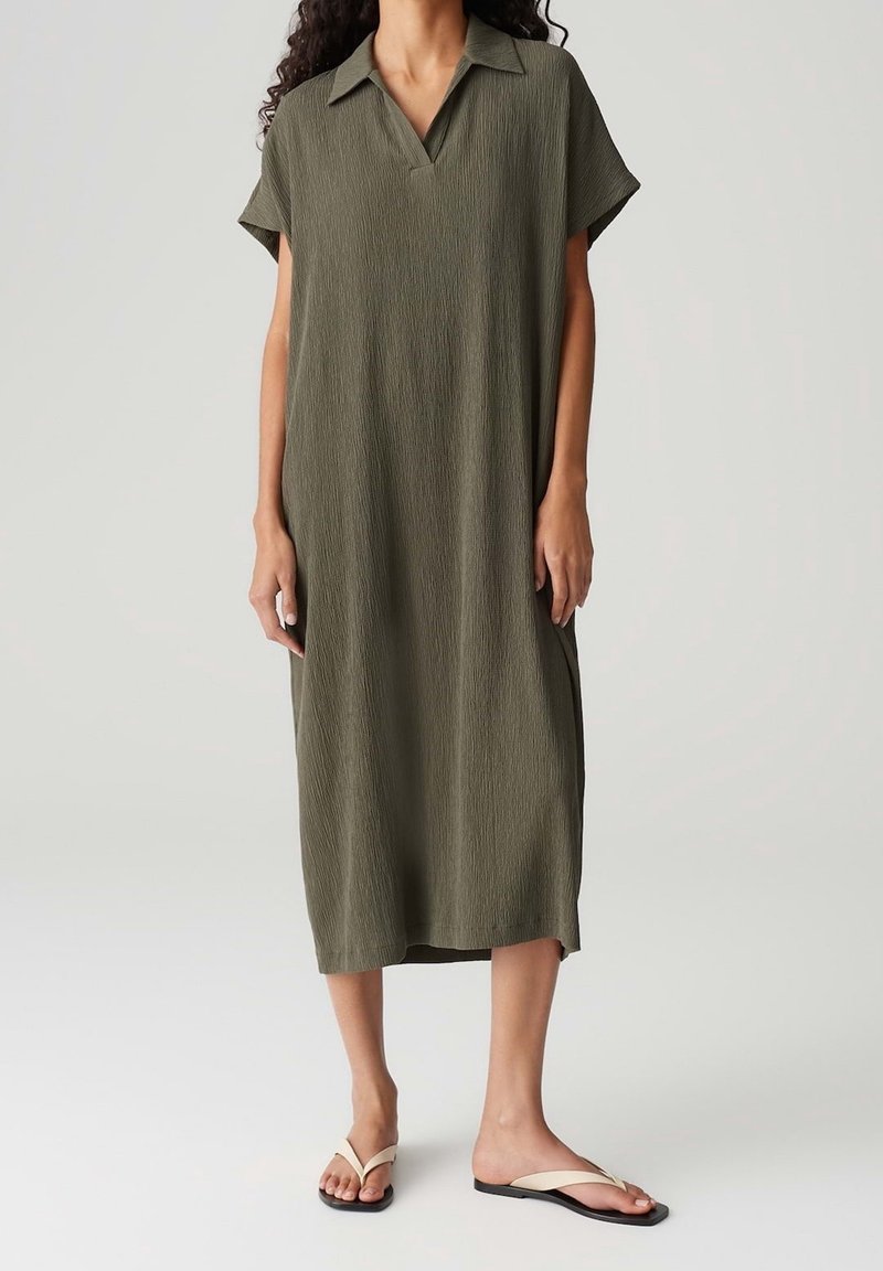 Femme portant une robe midi ample vert olive avec manches courtes et col, associée à des sandales plates blanches sur un fond uni.