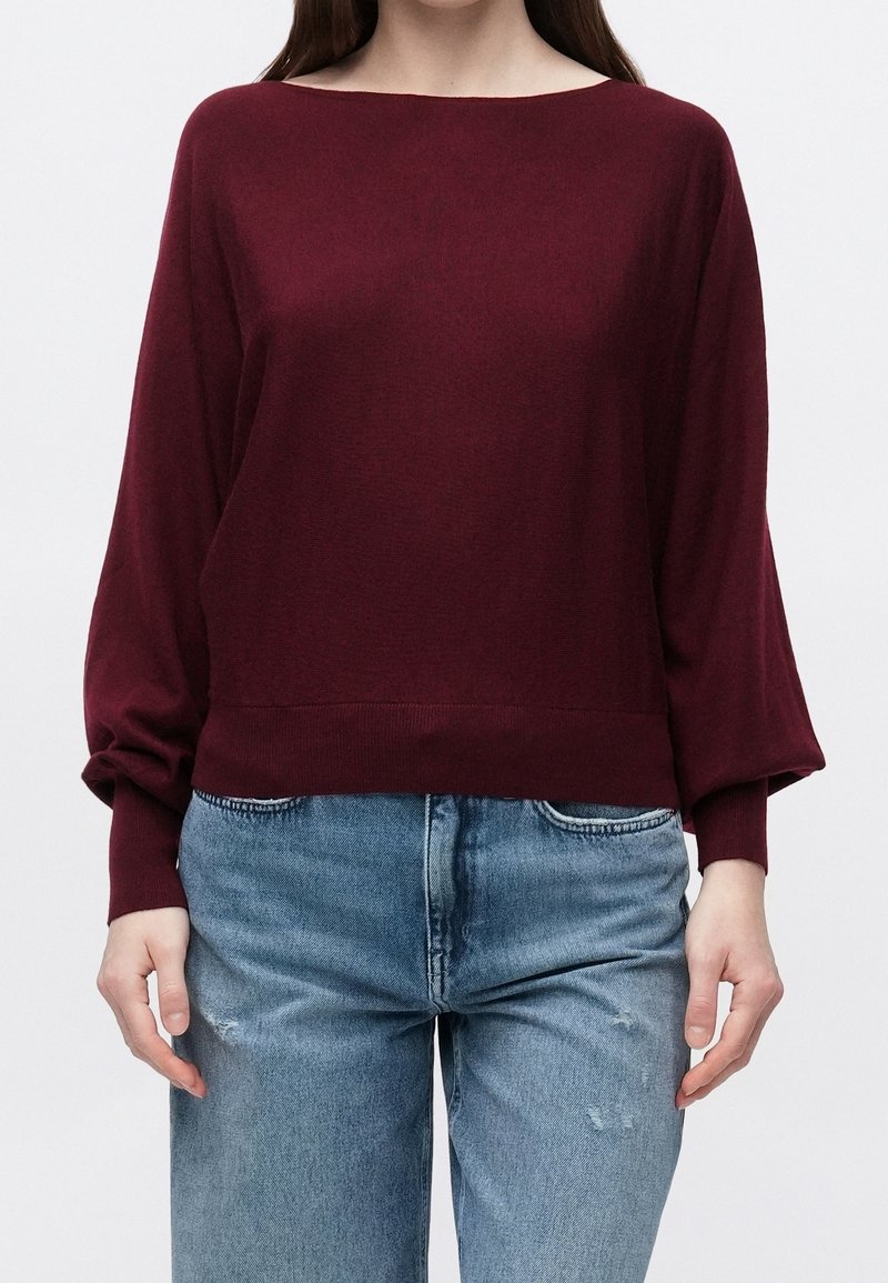 Femme portant un pull bordeaux à manches longues et col bateau, associé à un jean bleu clair déchiré, debout devant un fond blanc uni.