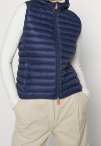 Gilet imbottito blu navy con cappuccio, caratterizzato da una cerniera frontale con tiretto arancione e cuciture diagonali per la texture. Vestibilità aderente.