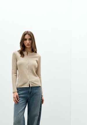 Jeune femme aux cheveux bruns porte un pull beige boutonné et un jean bleu, debout devant un fond blanc uni.