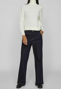 Pull à col roulé blanc associé à un pantalon large en denim foncé. Le pull présente une texture douce ; le pantalon possède des coutures apparentes et des poches.