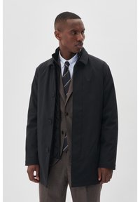 Cappotto impermeabile nero con chiusura a bottoni, colletto alto e tasche laterali, indossato sopra un gilet marrone e una camicia bianca con cravatta.