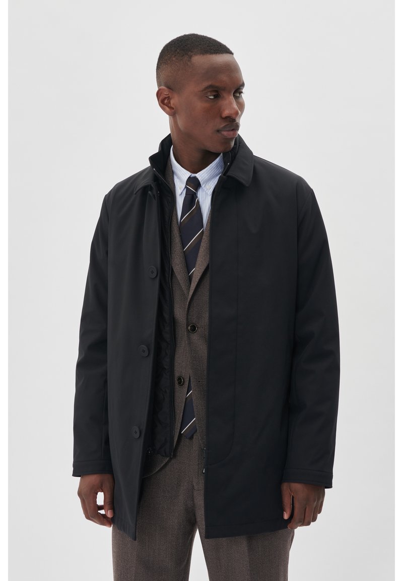Cappotto impermeabile nero con chiusura a bottoni, colletto alto e tasche laterali, indossato sopra un gilet marrone e una camicia bianca con cravatta.