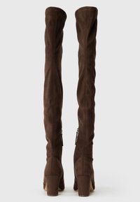 Par de botas altas de ante marrón con tacones de bloque y cremalleras laterales, mostradas desde atrás sobre un fondo claro y liso.