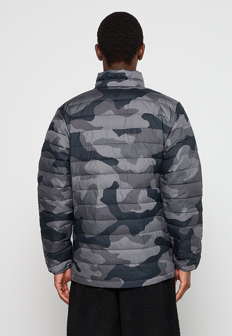 Hombre vestido con una chaqueta acolchada de camuflaje en tonos gris y negro y pantalones negros texturizados, visto desde atrás sobre un fondo blanco.