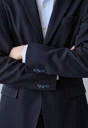 Person iført en mørk marineblå blazer over en lyseblå stribet skjorte, stående med armene over kors ved brystniveau.