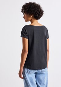 Černé tričko s krátkým rukávem a širokým výstřihem, vyrobené z měkké bavlny. Záda mají hladkou texturu, doplněné o světle modré džíny.