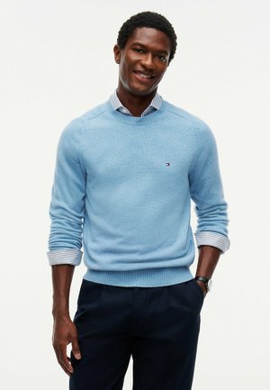Hombre sonriente con un suéter azul claro sobre una camisa a rayas con las mangas remangadas y pantalones oscuros, posando con una mano en el bolsillo.