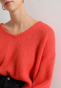 Maglione a maglia color corallo con scollo a V ampio e vestibilità ampia. Tessuto testurizzato con motivi di cuciture visibili, abbinato a jeans neri a vita alta.