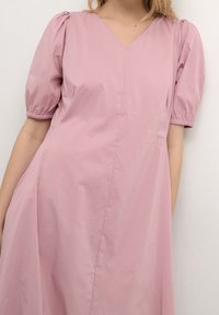 Rosa bomullsdräkt med V-ringning och korta puffärmar. Har en lös, svepande design med subtil sömdetalj och sidfickor.