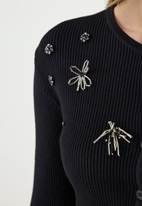Cardigan noir à côtes avec tissu texturé. Présente des embellissements en métal argenté en forme d'araignée et de ronds, enrichissant le design de formes et de détails.