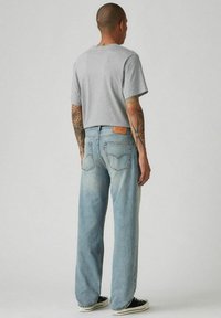 Hombre con tatuajes de espaldas, vistiendo una camiseta gris, jeans azul claro y zapatillas negras, frente a un fondo gris liso.