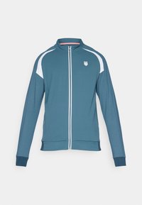 Jachetă teal cu fermoar, cu accente albe, mâneci lungi, guler înalt și un logo pe pieptul stâng. Textură netedă și design slim fit.