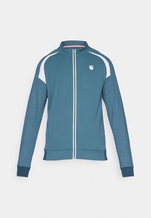 Teal värvi tõmblukuga jakk valgete aktsentidega, pika varrukaga, seisev kaelus ja logo vasakul rinnal. Sile tekstuur ja saledat sobivust rõhutav disain.