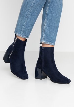 Personne portant des bottines en daim bleu à talons blocs et fermetures éclair latérales, associées à un jean en denim bleu clair.