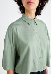 Blusa de manga curta verde claro com colarinho, apresentando um tecido texturizado e fecho de botões na frente. Design simples e ajuste relaxado com mangas oversized.