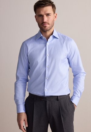 Mann mit hellblauem Hemd und dunkelgrauer Hose steht vor einfachem Hintergrund, eine Hand in der Tasche.