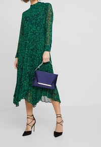 Robe à imprimé léopard vert avec manches longues et ourlet asymétrique fluide. Associée à une pochette marine et des talons noirs à brides.