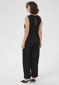 Tuta nera con design senza maniche, vita cintrata e dettaglio a occhiale sulla schiena. Tessuto testurizzato e pantaloni a gamba larga. Calzature nere.