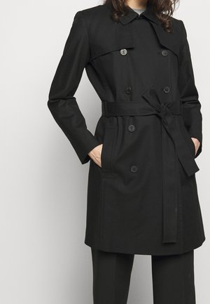 Vrouw draagt een zwarte dubbelgeknoopte trenchcoat met een ceintuur in de taille en handen in de zakken tegen een effen lichte achtergrond.