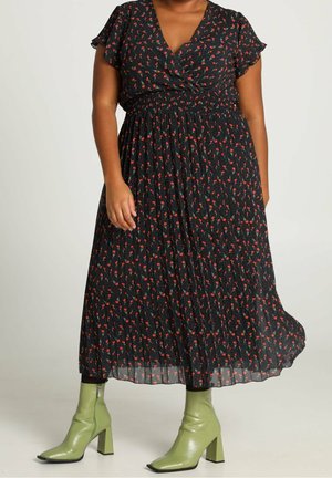 Kvinde iført en sort blomsterprintet midi-kjole med plisseringer og grønne hælede ankelstøvler, stående mod en ensfarvet lys baggrund.