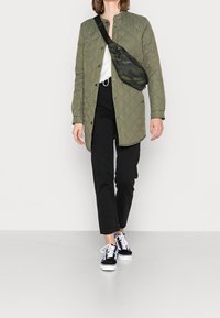 Persona vestida con un abrigo acolchado verde, pantalones negros, zapatillas negras y blancas, y una bolsa cruzada de camuflaje caminando frente a un fondo sencillo.
