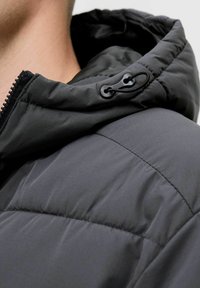 Schwarze gepolsterte Jacke mit verstellbarem Kapuze, strukturiertem Stoff und sichtbaren Nähten. Auch Reißverschluss und Hardwaredetails sind vorhanden.
