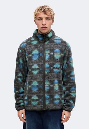 Jeune homme aux cheveux blonds courts portant une veste polaire grise avec des motifs circulaires bleus et verts et une fermeture éclair frontale.