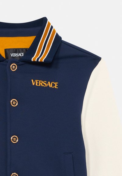 Tengerészkék sportdzseki fehér ujjal, arany gombokkal és narancssárga csíkos gallérral. Hímzett "VERSACE" logó található a mellrészen.