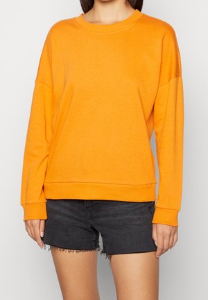 Personne portant un sweat-shirt à manches longues orange vif et un short en jean noir à ourlet effiloché, debout devant un fond uni.