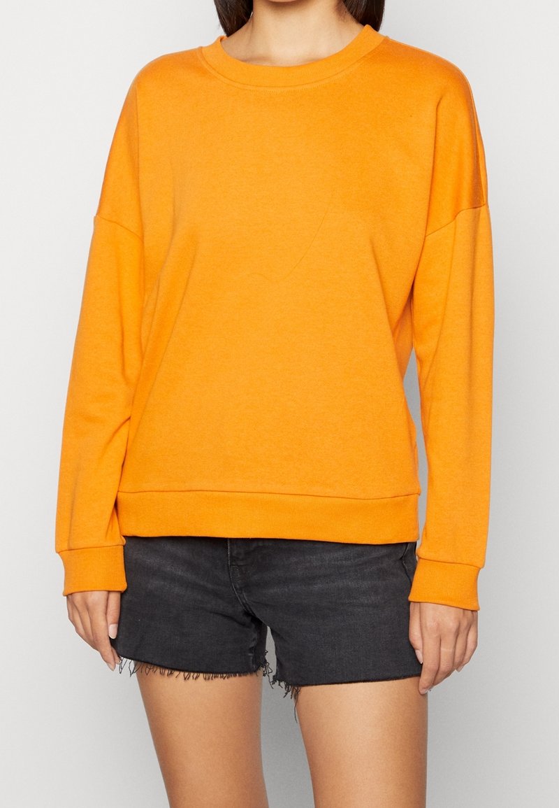 Personne portant un sweat-shirt à manches longues orange vif et un short en jean noir à ourlet effiloché, debout devant un fond uni.