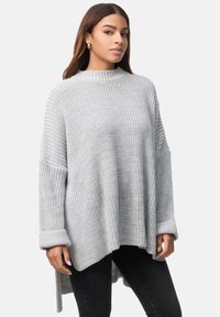 Hellgrauer gestrickter Pullover mit hohem Kragen, oversized Passform, weiten Ärmeln und gerippten Bündchen, kombiniert mit dunklen Jeans.