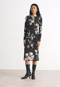Schwarzes Blumenmaxikleid mit langen Ärmeln und schmaler Silhouette, das aus einem transparenten Stoff mit weißen Blumenmustern besteht. Dazu passende schwarze kniehohe Stiefel.