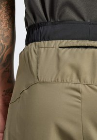 Pantalones cortos de color verde oliva hechos de una tela ligera con una cinturilla elástica negra. Presenta un bolsillo trasero con cremallera. Textura suave y diseño ajustado.