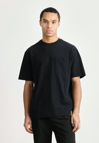 Μαύρο βαμβακερό t-shirt με στρογγυλή λαιμόκοψη, κοντά μανίκια και τσέπη στο στήθος. Διαθέτει μια μικρή πλευρική ετικέτα σε κίτρινο χρώμα. Απλή, άνετη εφαρμογή.