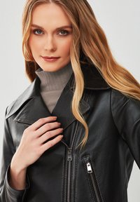 Zwart leren motorjack met grote revers, zilveren ritsen en zichtbare stiksels, gedragen over een lichtgrijze turtleneck.