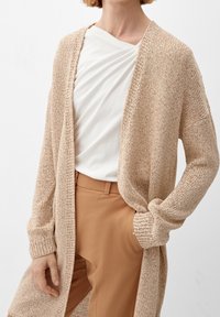 Cardigan beige lavorato a maglia con apertura frontale e maniche lunghe, abbinato a una maglietta bianca e pantaloni beige. Il tessuto ha un aspetto strutturato.