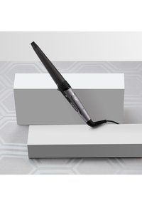 Svart krølltang med en konisk sylinder, metallisk sølvhåndtak og LED-kontroller, som hviler på hvite rektangulære blokker mot en teksturert overflate.