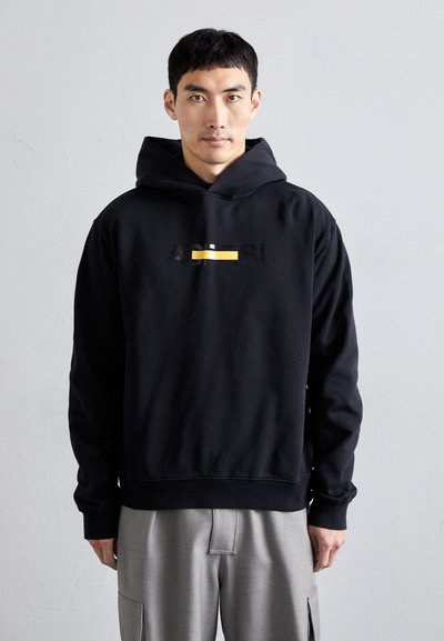 Black hooded sweatshirt with a front graphic in black and yellow. Features a relaxed fit and ribbed cuffs. Worn with light grey trousers.