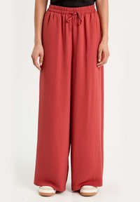 Un pantalon large en tissu texturé rouge brique, doté d'une taille élastique avec un cordon de serrage. Associé à des baskets beiges.