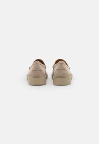 Semišové slip-on boty v světlé béžové barvě s kulatými okraji, prošitými detaily a hnědým interiérem, umístěné na béžové gumové podrážce.