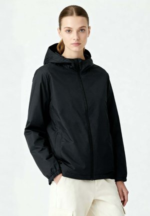 Vrouw met een lichte huid en naar achteren gebonden haar draagt een zwart gewatteerd jack met capuchon en crèmekleurige cargobroek, staand tegen een effen lichte achtergrond.