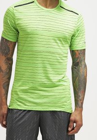 Camisa atlética de color verde neón con patrones de olas horizontales, mangas cortas, cuello redondo y detalles en negro en los hombros. Llevada con pantalones cortos con patrones.