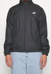 Svart Nike vindjacka med hög krage, dragkedja framtill, elastiska ärmslut och en liten vit logga på vänster bröst. Slätt syntetmaterial.
