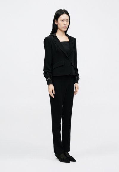 Mustast kärbitud kapuutsiga jakk, kehatut must top ja lõigatud püksid. Jakil on tekstureeritud mansetid ja kerge õlapadi.