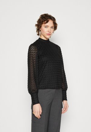 Black sheer blouse with textured pattern, high neck, and long sleeves. Paired with gray tailored pants, showcasing a formal look.