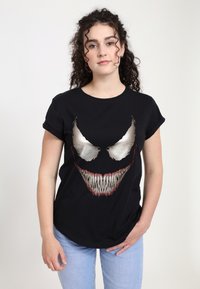 Sort bomulds T-shirt med korte ærmer, der har en stor, stiliseret grafik af et udtryksfuldt ansigt med overdrevede tænder og øjne.